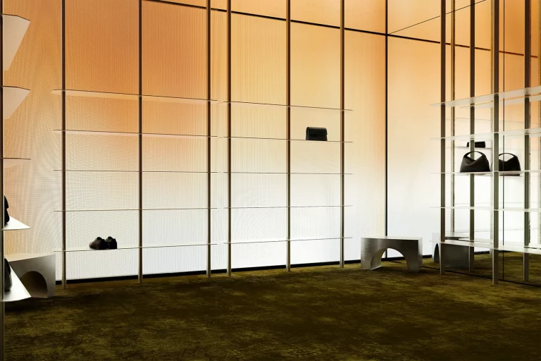 NOVU flagship retail concept by NOCOD Studio — full-height backlit amber LED panels, transparent glass shelving, brushed steel arched bench, deep olive carpet. Display gallery.