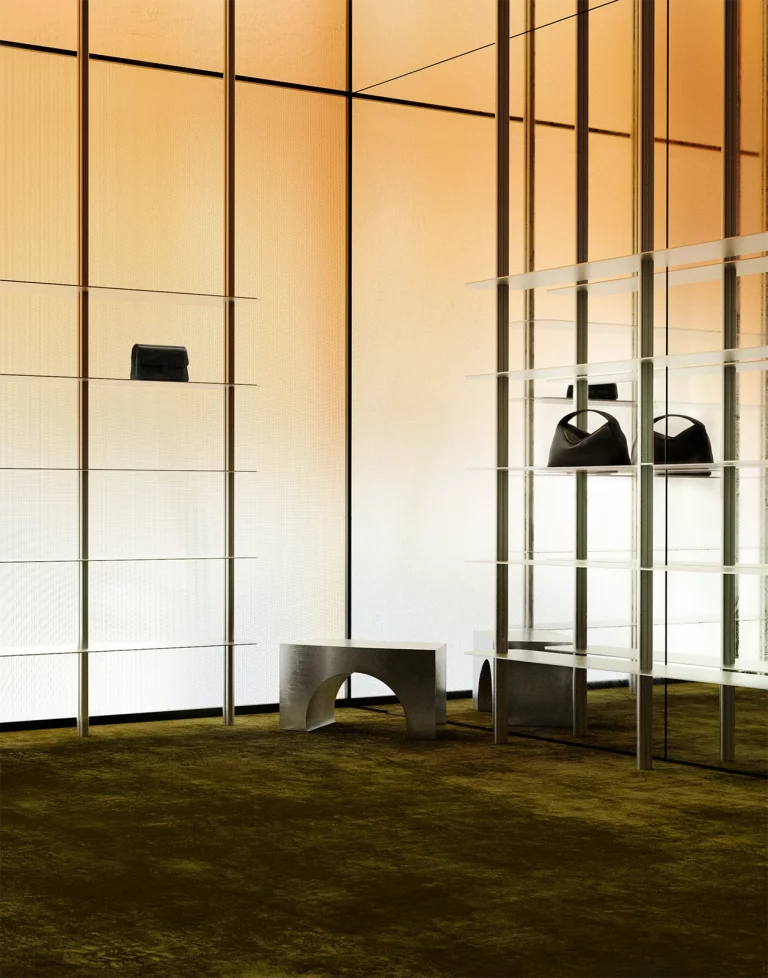 NOVU concept store retail gallery — amber LED panel wall, transparent shelving system, brushed steel arched bench, deep olive carpet. NOCOD Studio.