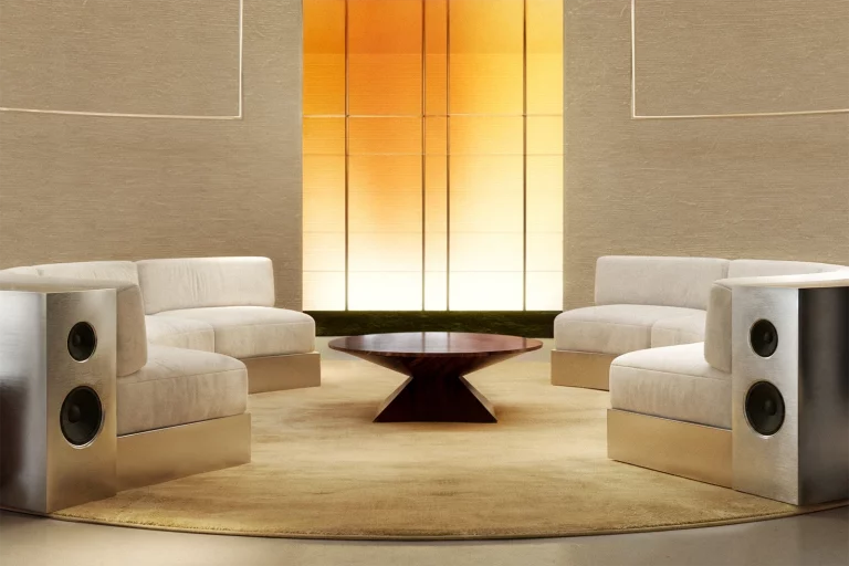 NOVU central listening salon by NOCOD Studio — two curved sofas with integrated speaker panels facing symmetrically. Round dark wood coffee table, amber backlit wall, cream silk carpet.